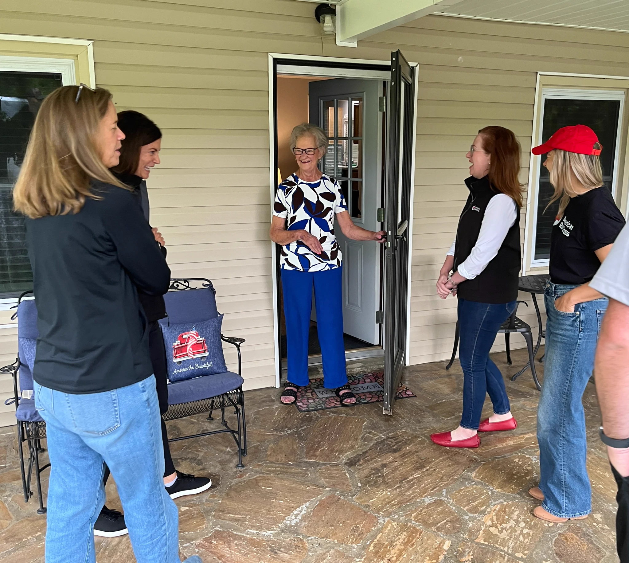 A person stands in an open doorway, smiling and greeting a group of visitors on a porch. The group includes five people, some standing and others sitting on a bench with a red hat visible. All are casually dressed.
