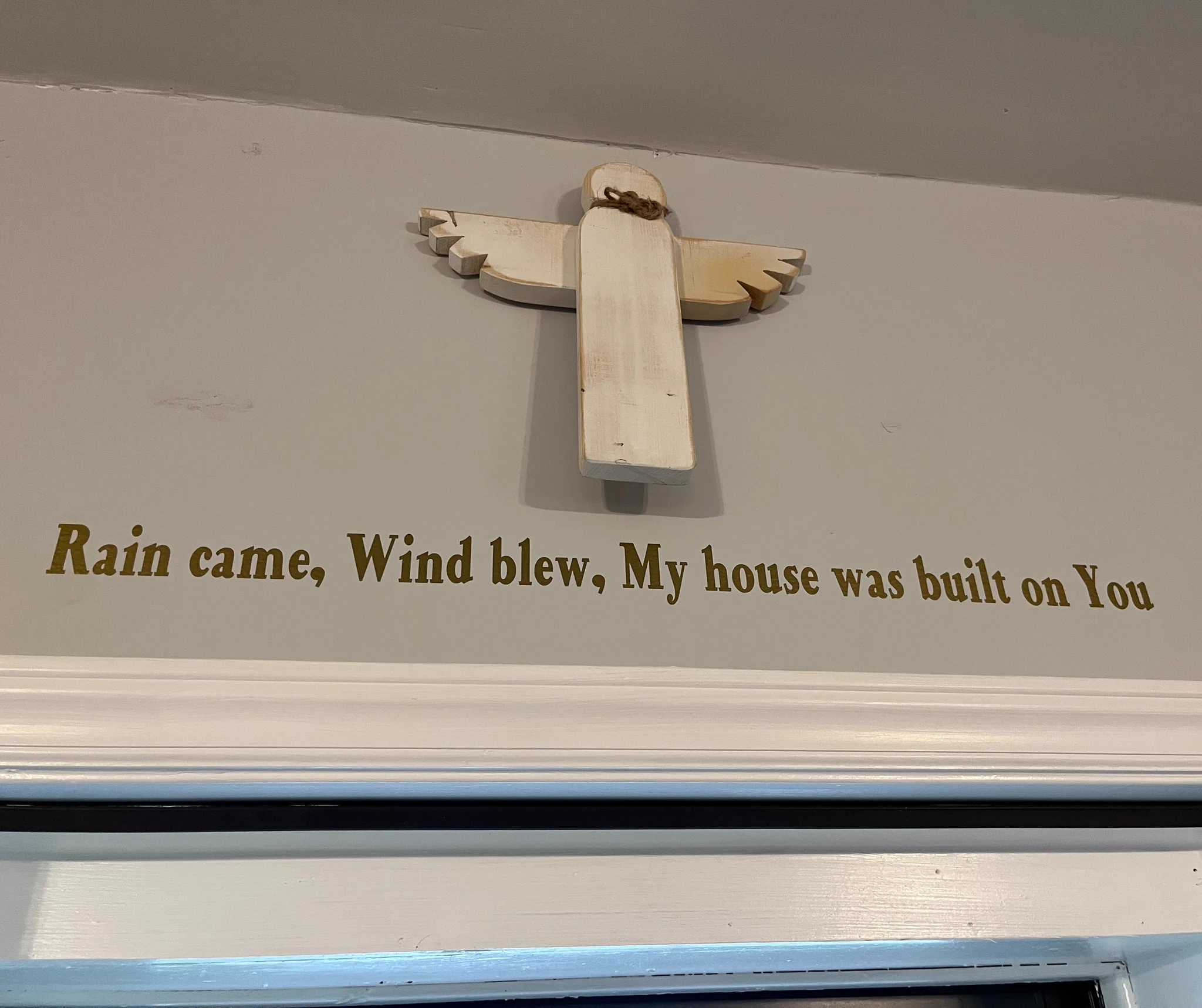 A wooden angel figure is mounted on the wall above a doorframe. Below it, text reads: "Rain came, Wind blew, My house was built on You.