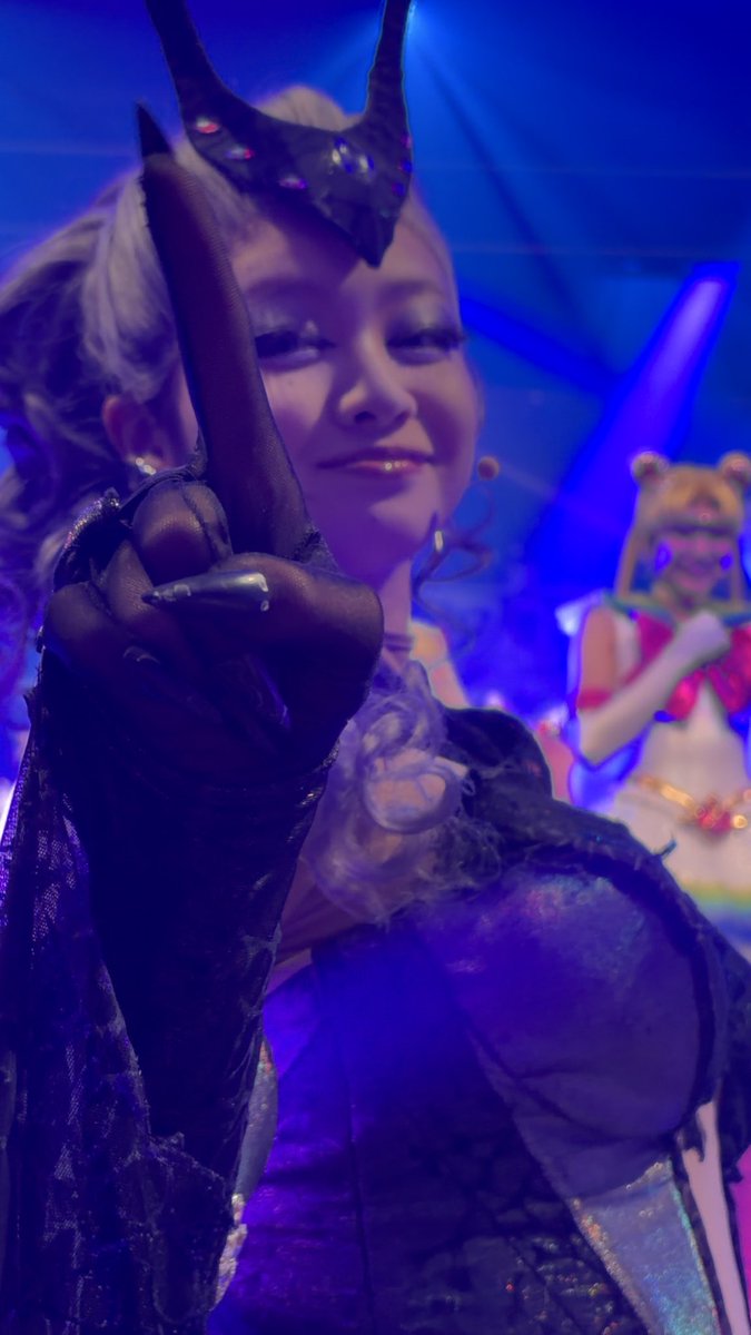 A cosplayer with blonde hair and a horned headpiece smiles warmly while posing for the camera, dressed in a dark purple costume with elaborate gloves, likely portraying the character Sakura Valkyrie. The image captures the performer's joyful fan service moment, matching the post's note about the idol looking delighted while pointing at the photographer during a live event.