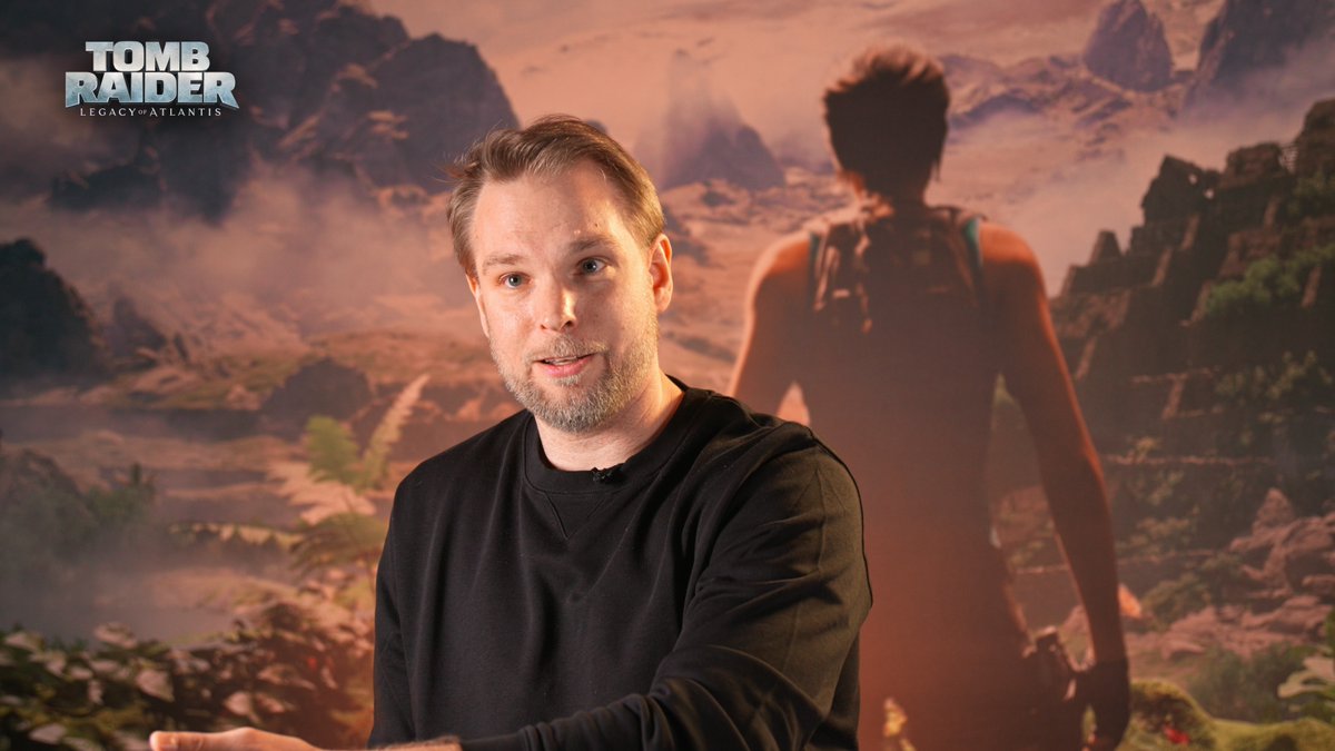 A bearded man in a black sweatshirt speaks during an interview, with the 'TOMB RAIDER LEGACY OF ATLANTIS' game logo in the corner and a screenshot of the game showing a female character in a misty landscape behind him.