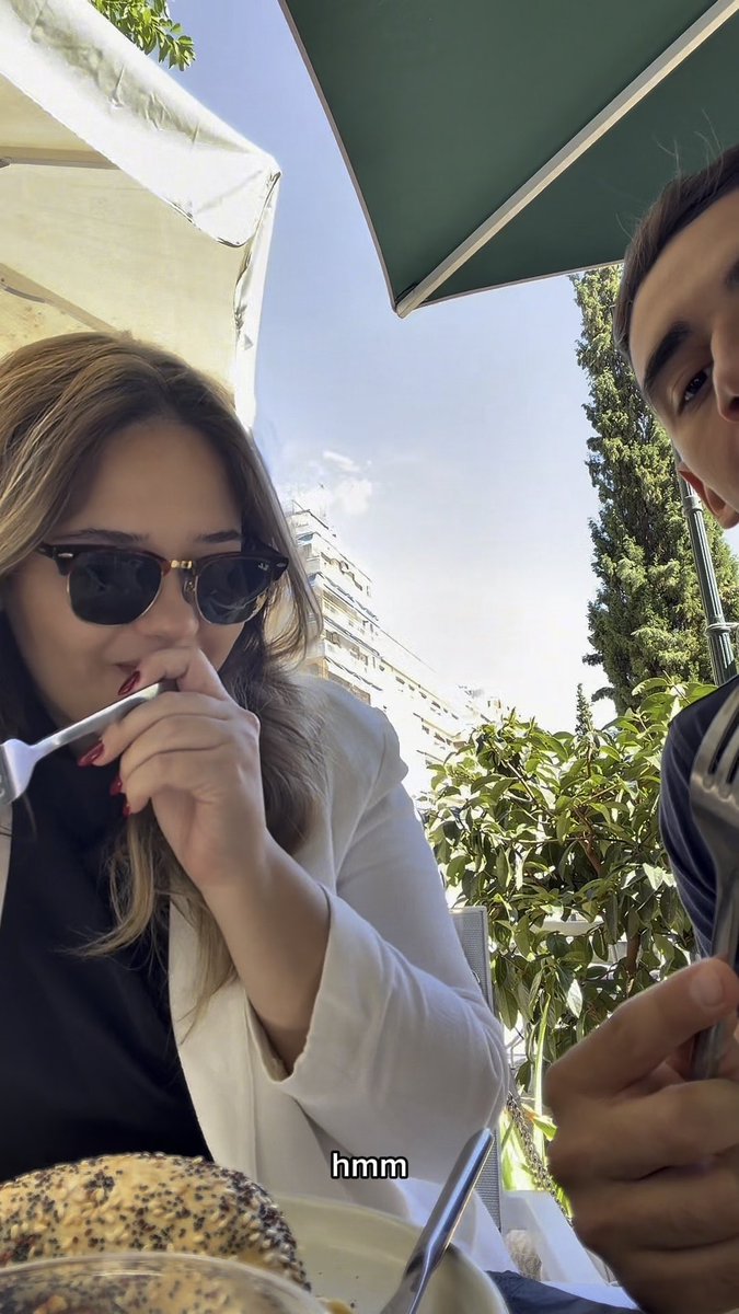 Two people sit at an outdoor cafe table under umbrellas, with a woman wearing sunglasses eating with a fork while a man looks on; the word 'hmm' is visible as text overlay.