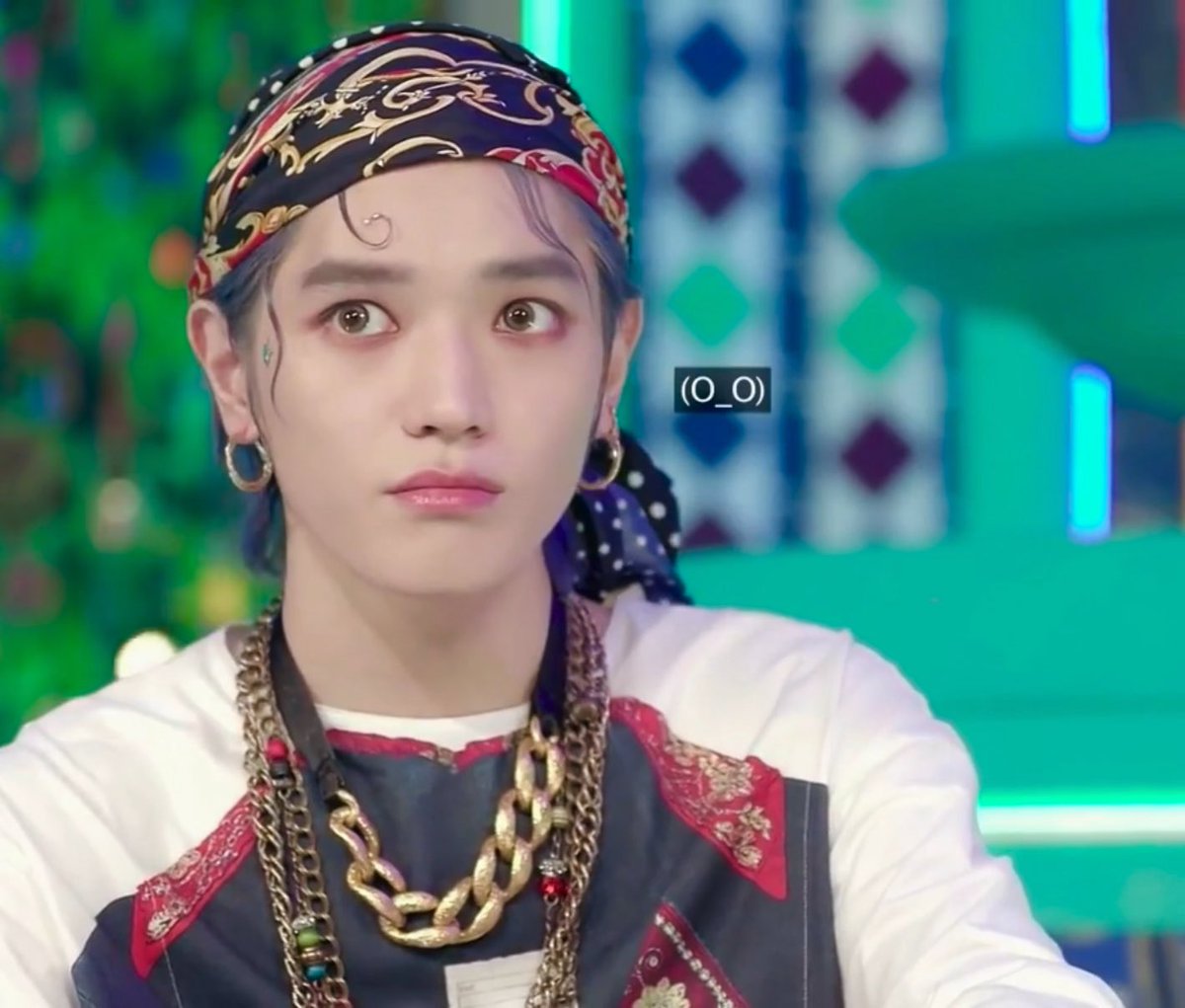 A young man with dark hair wearing a patterned bandana, gold hoop earrings, and layered gold chains makes a surprised facial expression with the text '(O_O)' overlaid next to him.