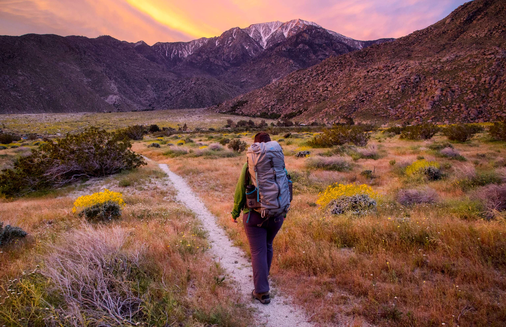 Hiker walking along a narrow trail through a grassy valley with mountains and a colorful sunset sky in the background.