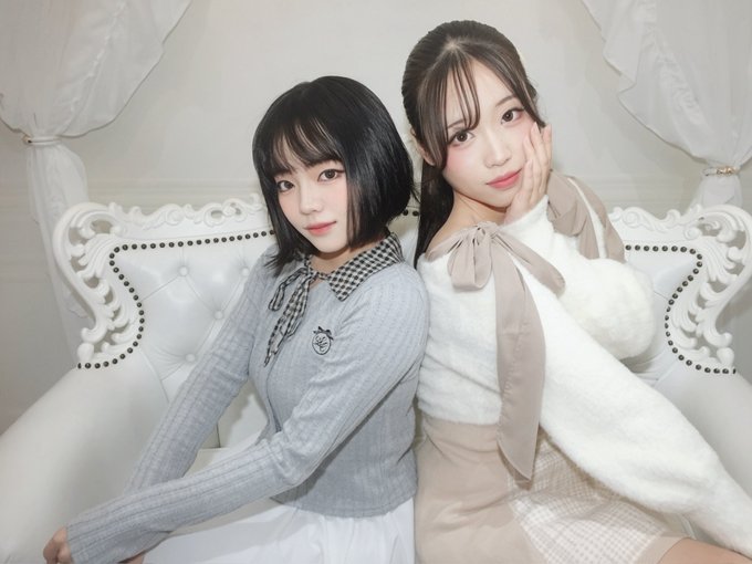 Two young women pose together on a white ornate couch; one wears a gray sweater with a checkered collar and the other wears a cream-colored top with a tan ribbon.