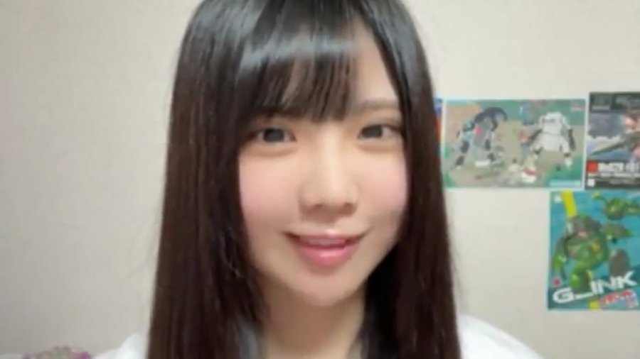 A smiling young woman with long black hair and bangs stands in front of a wall with gaming posters, including one for a 'G-LINK' game.