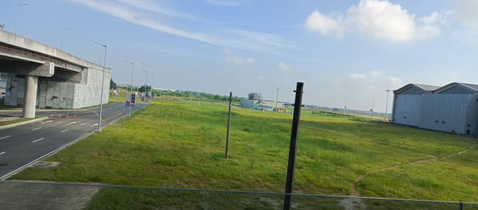 A serene morning view shows a grassy field beside a quiet road with an overpass on the left and a large warehouse on the right under a partly cloudy sky. The tranquil landscape perfectly complements the message's invitation to pause and appreciate the peaceful beauty of the moment.