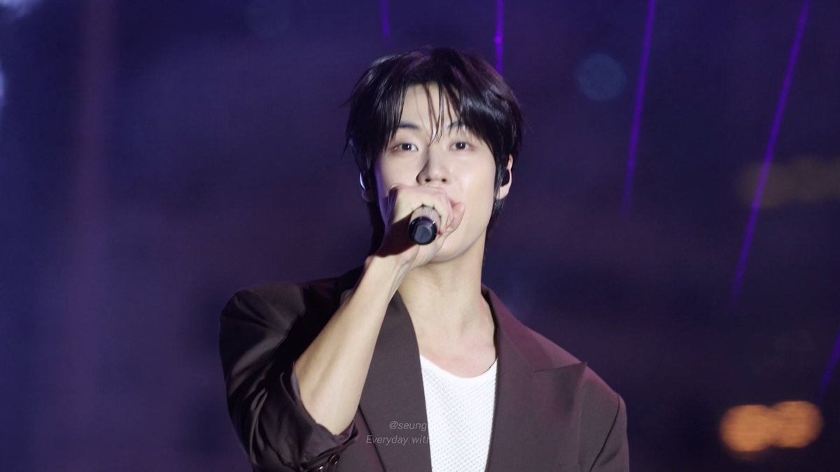 A young male performer sings into a microphone on stage under purple lighting, wearing a brown blazer over a white top, with a watermark reading "@seung Everyday with".