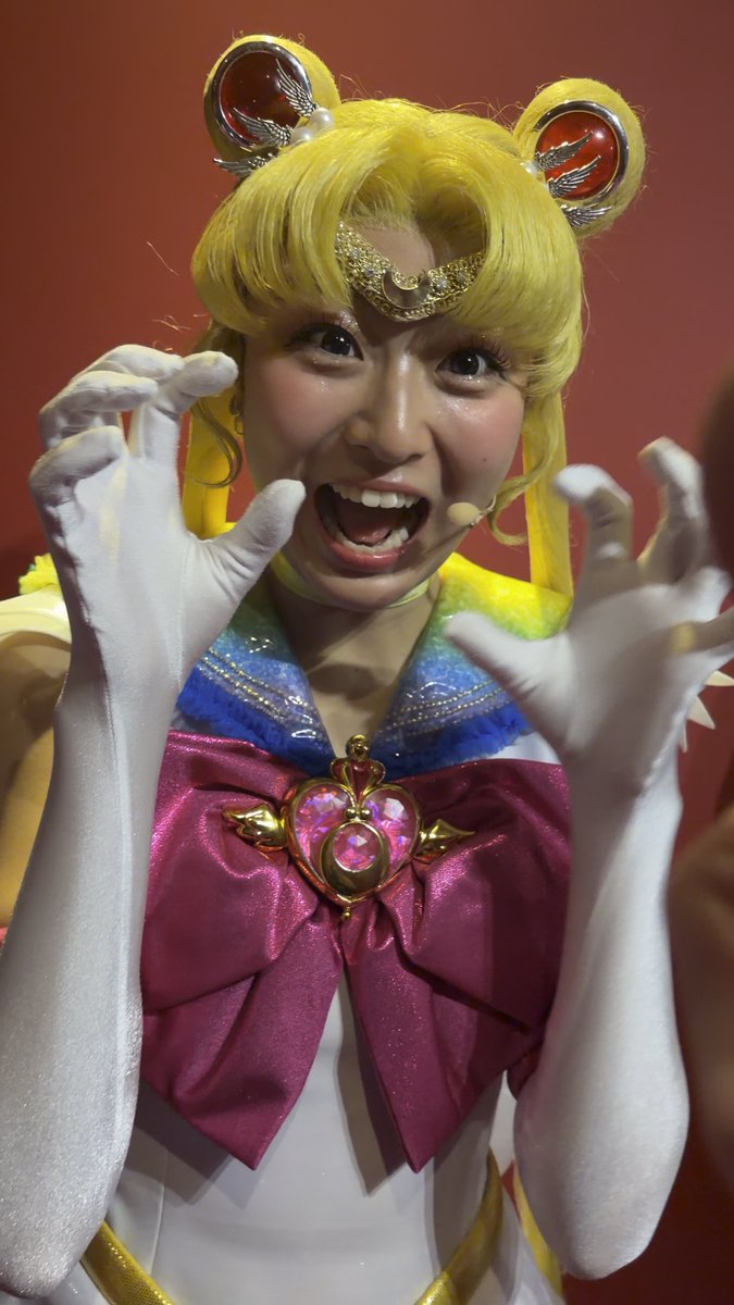 A performer in a Sailor Moon cosplay costume with blonde hair, a tiara, and a pink bow, posing dramatically with white-gloved hands raised near her face.