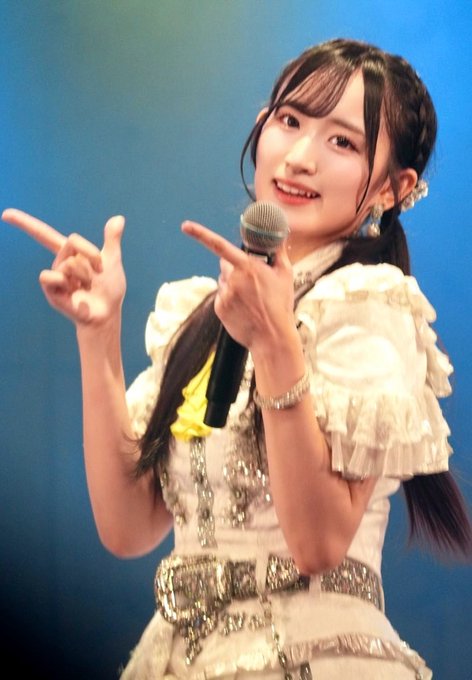 A young woman with long dark hair holds a microphone and gestures with both hands while wearing a white ruffled outfit with silver embellishments against a blue background.