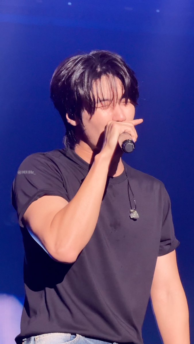 A photo of a young male performer with dark hair singing into a microphone on stage, wearing a black t-shirt and a pendant necklace, with the watermark '@1031Hz_'.