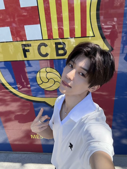 A young man in a white polo shirt takes a selfie in front of an FC Barcelona crest, with the visible text "FCB" and "MÉS".