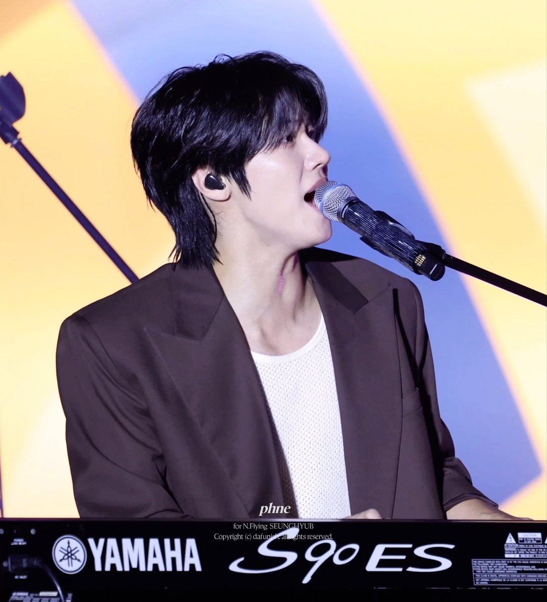 A male performer with black hair sings into a microphone while playing a Yamaha S90 ES keyboard, wearing a brown blazer over a white shirt.