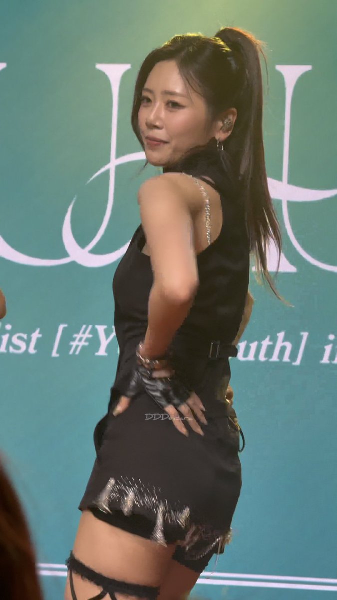 A female performer on stage wearing a black outfit with a high ponytail, posing in front of a teal backdrop with partial text visible including 'ist' and 'uth'.