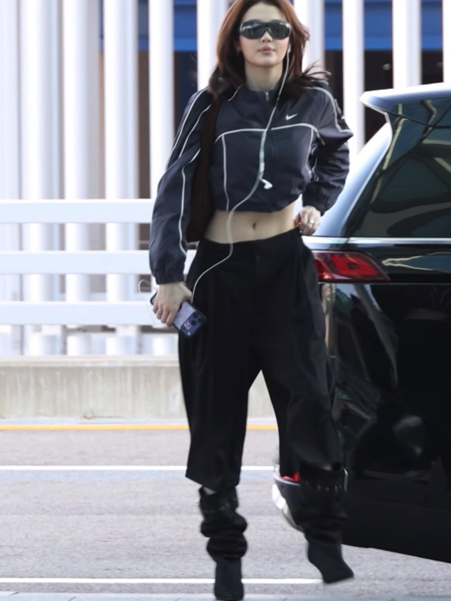 A woman wearing a black Nike crop jacket, black pants, and sunglasses walks through a parking lot next to a black car.