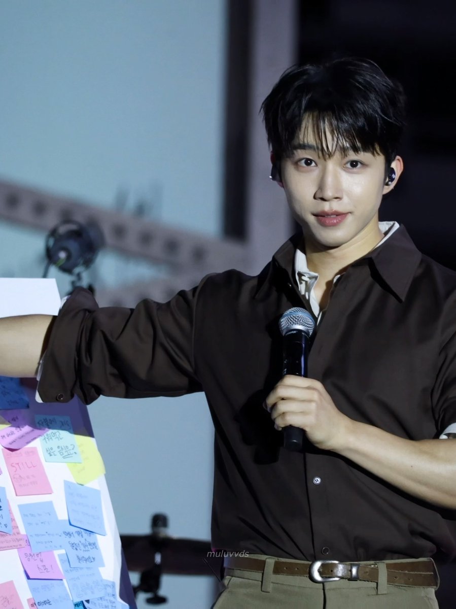 A young man with dark hair wearing a brown shirt speaks into a microphone while holding a board covered in colorful sticky notes with writing on them, with a watermark 'muluvvds'.