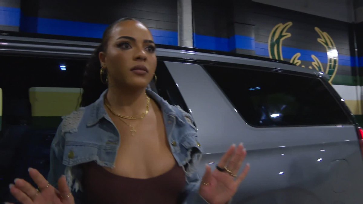 A woman with long dark hair, wearing a denim jacket over a brown top with layered gold necklaces, gestures with her hands while speaking in front of a silver SUV at what appears to be a garage or studio with branded signage. The image relates to the post as it likely captures a moment during the live post-show reaction on SiriusXM's WWE channel, possibly featuring Tommy's guest appearance at an international tour event.