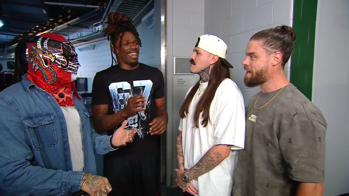 Four men, one wearing an ornate red and gold luchador mask and denim jacket, stand backstage in a hallway with a "COURTESY FACILITIES" sign visible on the door behind them, suggesting a WWE backstage encounter involving Je'Von Evans.