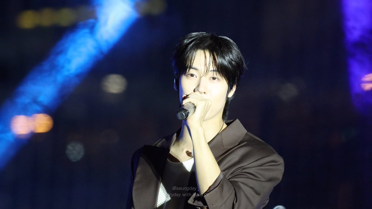 A young male performer sings into a microphone on stage under blue lighting, wearing a dark suit with a watermark reading '@seungday_everyday with #iKON'.