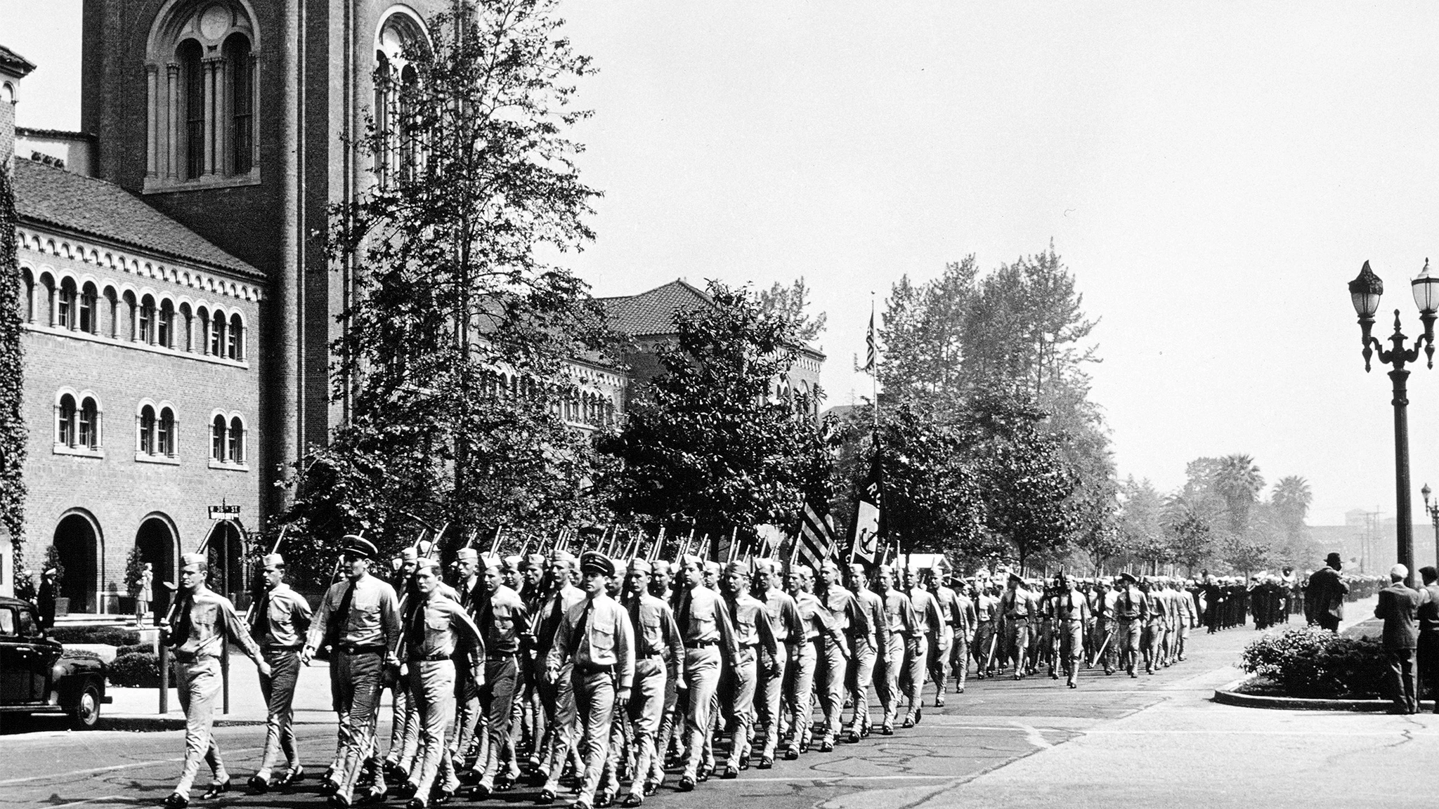 Black-and-white archival photo of uniformed USC military cadets marching in formation along a tree-lined street beside campus buildings, carrying rifles and flags.