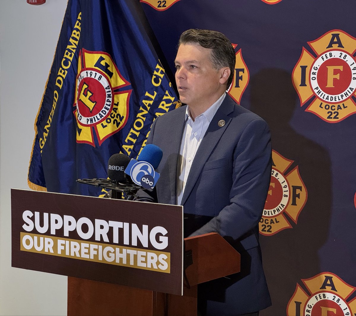 A man in a suit speaks at a wooden podium with several microphones in front of firefighter union flags and emblems. The podium displays a sign, and additional text appears on the flags and backdrop.
