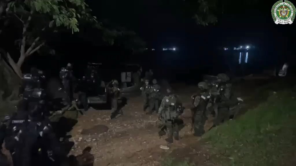 A nighttime image showing a group of armed military or police personnel in tactical gear gathered near a vehicle and a body of water, with a government emblem in the top right corner.