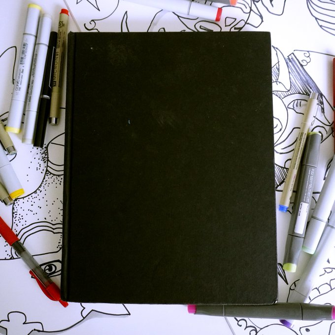 A closed black sketchbook lies flat on a surface decorated with black-and-white line art, surrounded by various pens and markers.