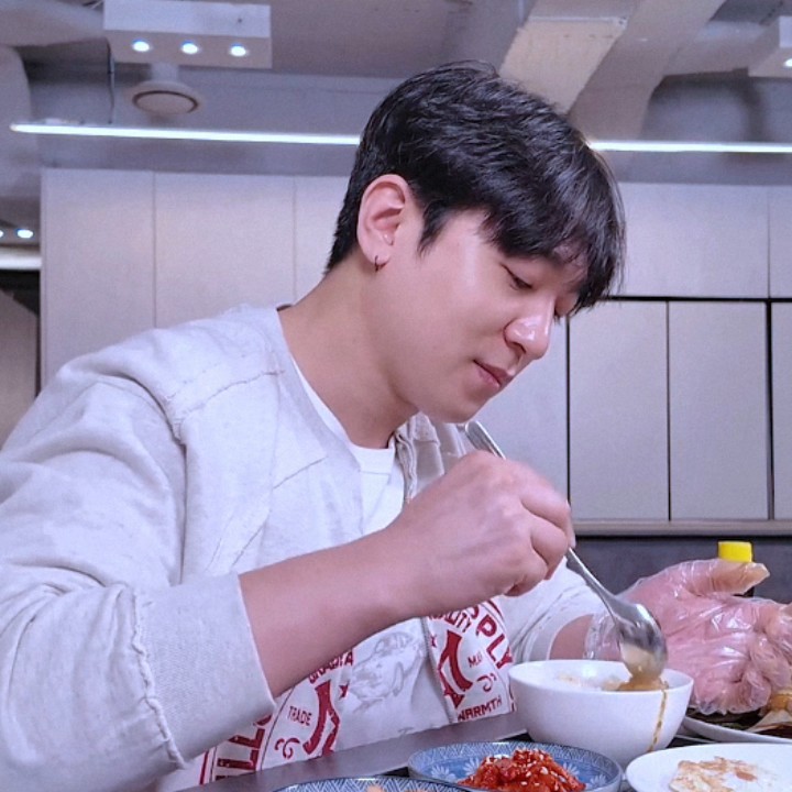 A man in a grey hoodie and a graphic apron eats a meal with a spoon, with a side of kimchi visible on the table.