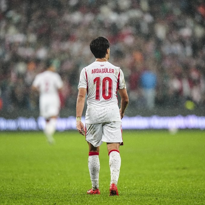 A soccer player wearing a white jersey with 'ARDAĞÜLER 10' on the back stands on a wet grass field, looking toward the blurred stadium crowd.