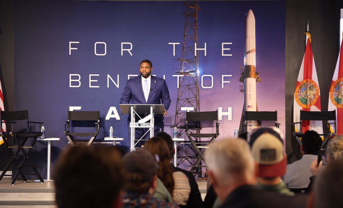 A man in a suit stands at a lectern on a stage before a large backdrop showing a rocket and launch tower. Empty chairs and small tables are arranged onstage, with an audience in the foreground and flags at the sides. There is text on the backdrop and lectern.