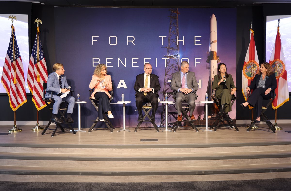 A panel discussion on a stage, with six people seated in chairs in front of a backdrop featuring a rocket and launch tower. Several flags stand on either side, and there is large text on the backdrop.