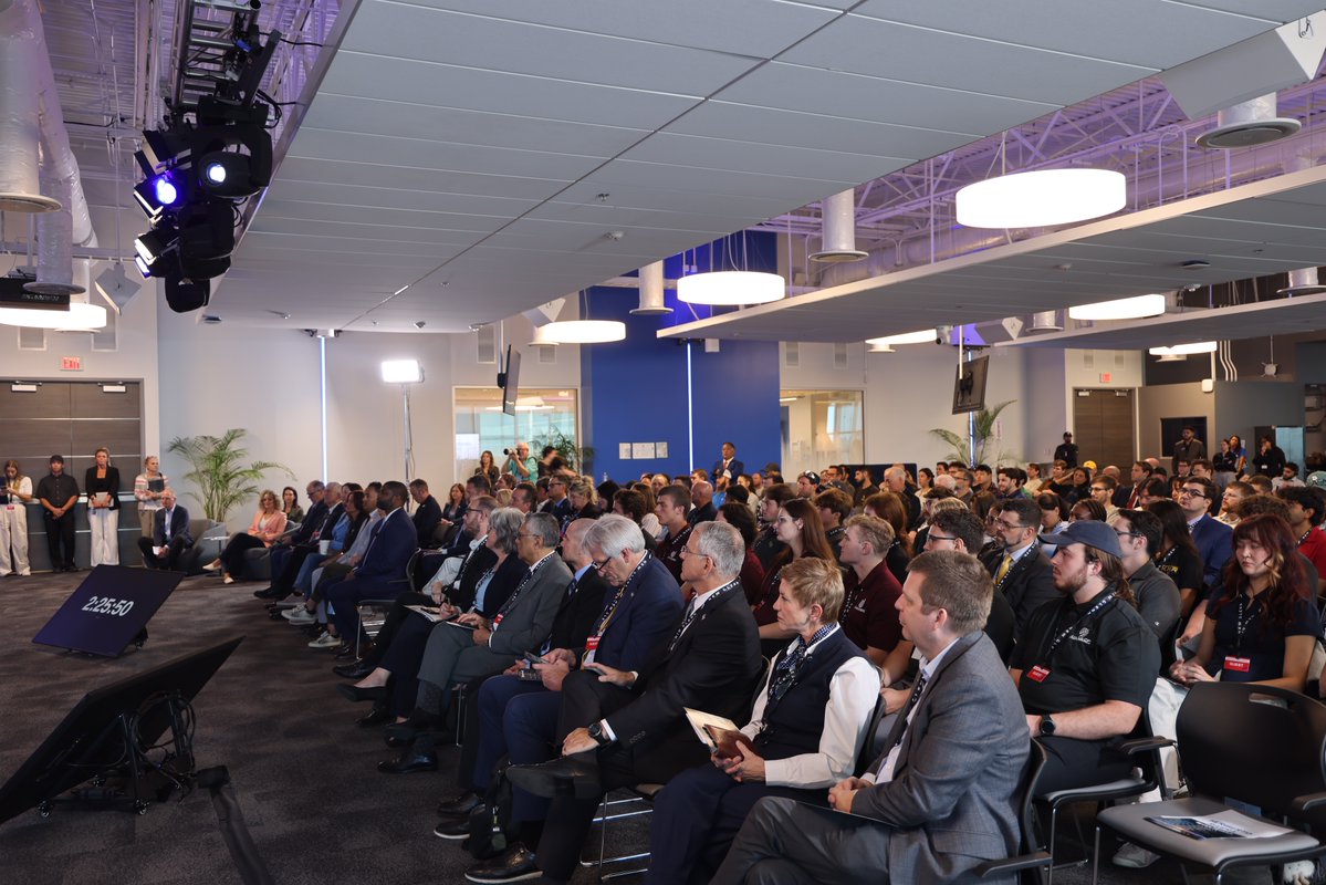 A large audience sits in rows at an indoor presentation or conference. Stage lighting and equipment are visible at the left, and a floor monitor displays text.