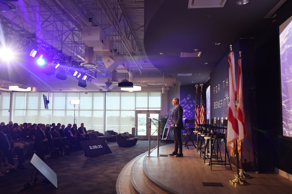 A man stands at a clear lectern on a stage, addressing an audience seated in a modern event space. Stage lighting, flags, chairs, and large screens are visible. There is text on the wall and a timer display.