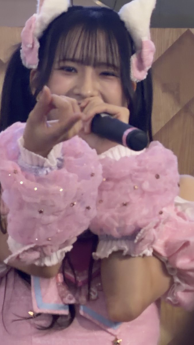 A close-up of a smiling idol wearing fluffy pink cat-ear headbands and a pink frilly outfit, holding a microphone close to her face.