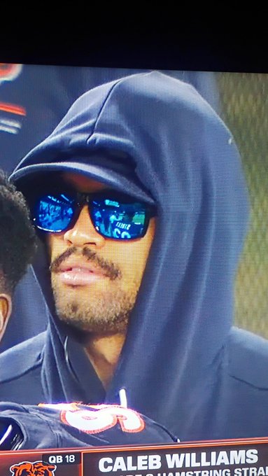 Close-up of a bearded man wearing a dark hoodie, cap, and blue reflective sunglasses; the sports broadcast graphic reads 'CALEB WILLIAMS' and 'QB 18'.