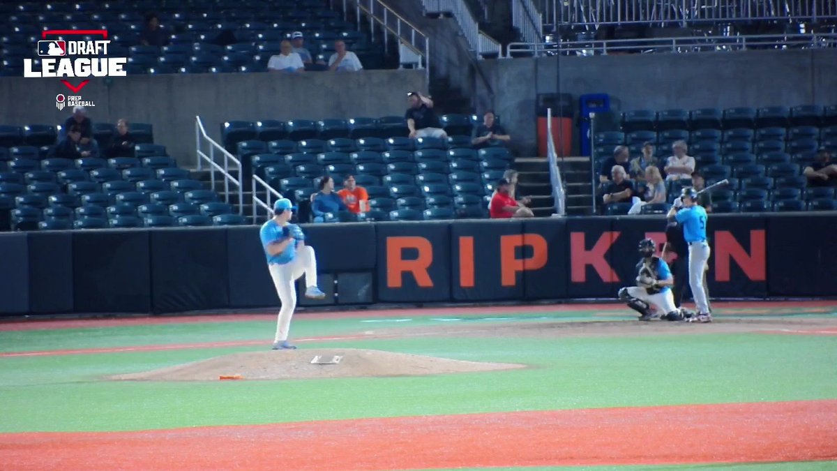 A pitcher in a light blue jersey winds up on the mound while a batter stands ready at home plate during a baseball game, with 'DRAFT LEAGUE' and 'RIPKEN' signage visible.