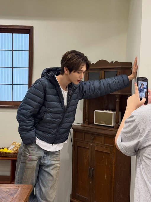 A young man with face tattoos wearing a blue puffer jacket and jeans poses against a wall while someone off-camera takes his photo with a smartphone.
