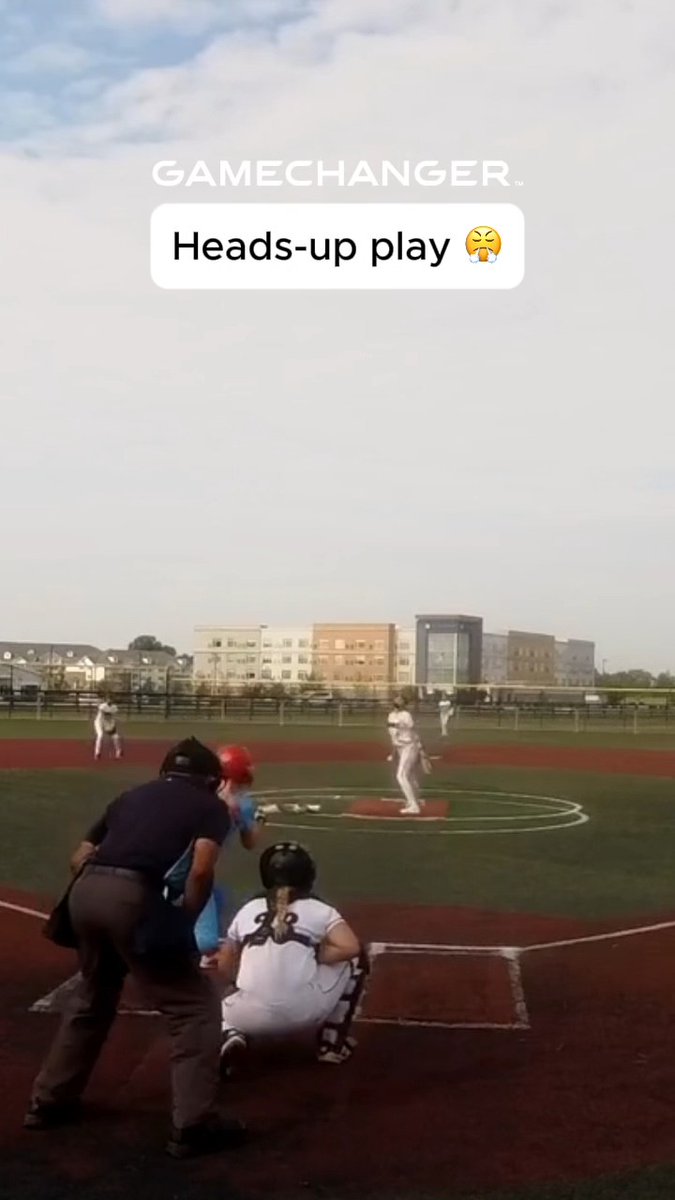 A GameChanger app screenshot showing a softball game at bat with the text "Heads-up play 😩" overlaid on a cloudy sky view of a softball field with players visible.