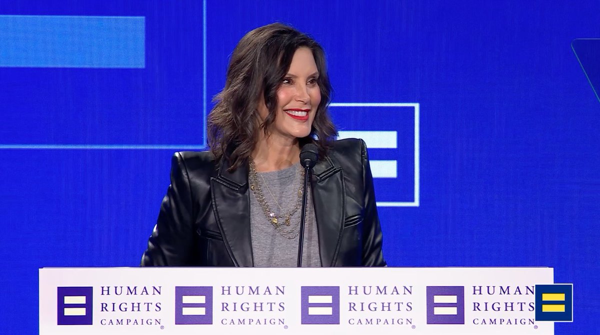 A woman with dark wavy hair wearing a black leather blazer and red lipstick smiles at a microphone on stage at a Human Rights Campaign event.