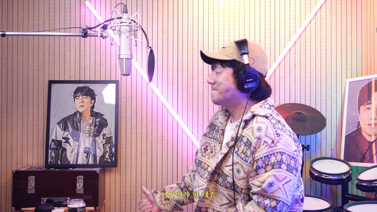 A man wearing a tan cap and headphones stands in a recording studio near a microphone, with a portrait painting visible and Korean subtitles "낭만이라 할 테죠" displayed.