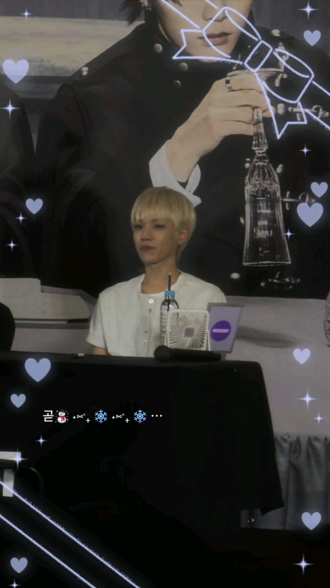 A person with short blonde hair wearing a white shirt sits at a table with a water bottle and small fan, in front of a large promotional poster, with heart graphics and Korean text '곰🐻✿+❄❄+❄...' overlaid.