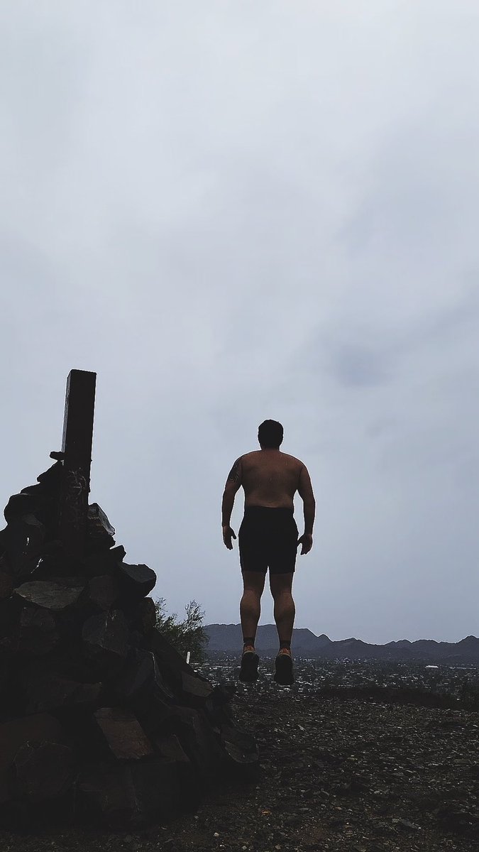 A silhouette of a shirtless man in shorts standing on a rocky hilltop, viewed from behind against an overcast sky with mountains in the distance.