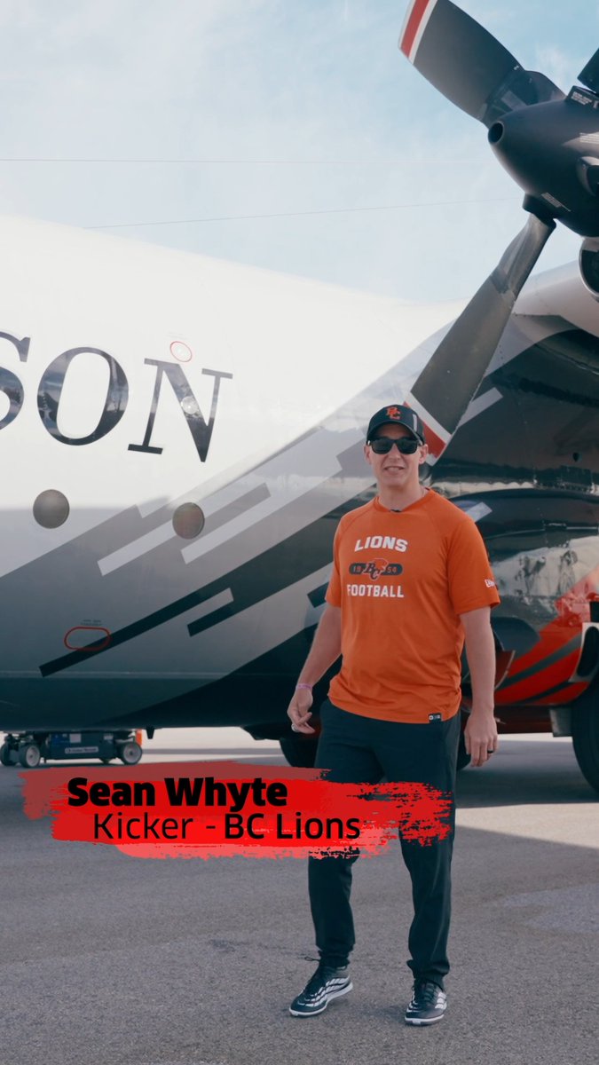 BC Lions kicker Sean Whyte stands smiling in front of a team-branded airplane while wearing an orange 'Lions Football' shirt, black sweatpants, and a baseball cap.