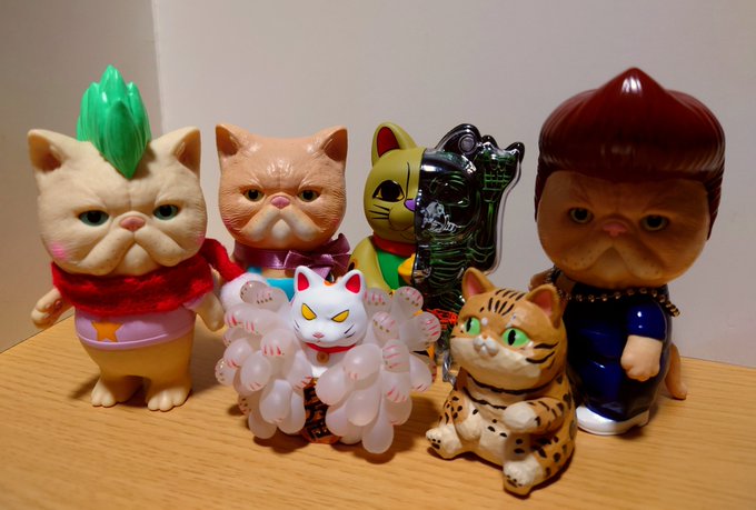 A collection of cat figurines sits on a wooden surface, including figures resembling a green-haired cat, a cat with multiple ghost paws, and a small tiger cat.