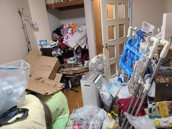 A cluttered, hoarded room is filled with piles of clothing, cardboard boxes, plastic bags stuffed with assorted items, a fan, and PVC pipes stacked haphazardly by the doorway. The image illustrates the cautionary point in the post: when someone tries to save money by packing their own bags, the result is often overstuffed, disorganized bags where everything is jumbled together rather than sorted.
