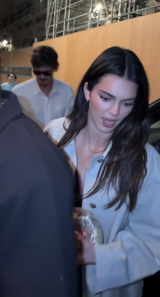 A woman with long dark hair wearing a light blue button-up shirt appears to be looking down, possibly signing autographs or handling an item, while a man in a white shirt and sunglasses stands behind her near scaffolding in what looks like an outdoor Parisian setting. The image captures Kendall Jenner arriving at a Paris restaurant, consistent with the post about her and Jacob Elordi's appearance in France on September 28.