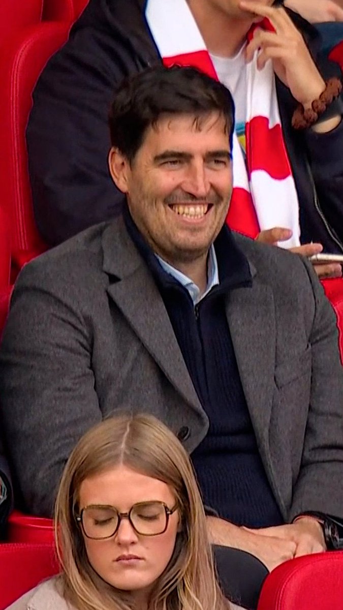 Andoni Iraola, the Bournemouth men's first team manager, smiles broadly while watching from the stands at Anfield during the Women's Super League Merseyside derby between Liverpool and Everton, which Liverpool won 2-0. He is seated among spectators wearing a grey coat over a navy zip-up and blue shirt.