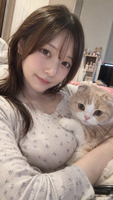 A young woman with long brown hair wearing a white floral top smiles at the camera while holding a chubby orange and white Scottish Fold cat.