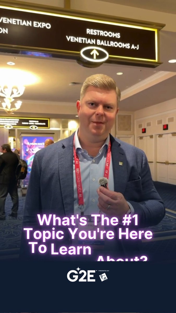 A man in a blue blazer at a conference holds a microphone, with overlay text reading 'What's The #1 Topic You're Here To Learn About?' and a G2E logo.