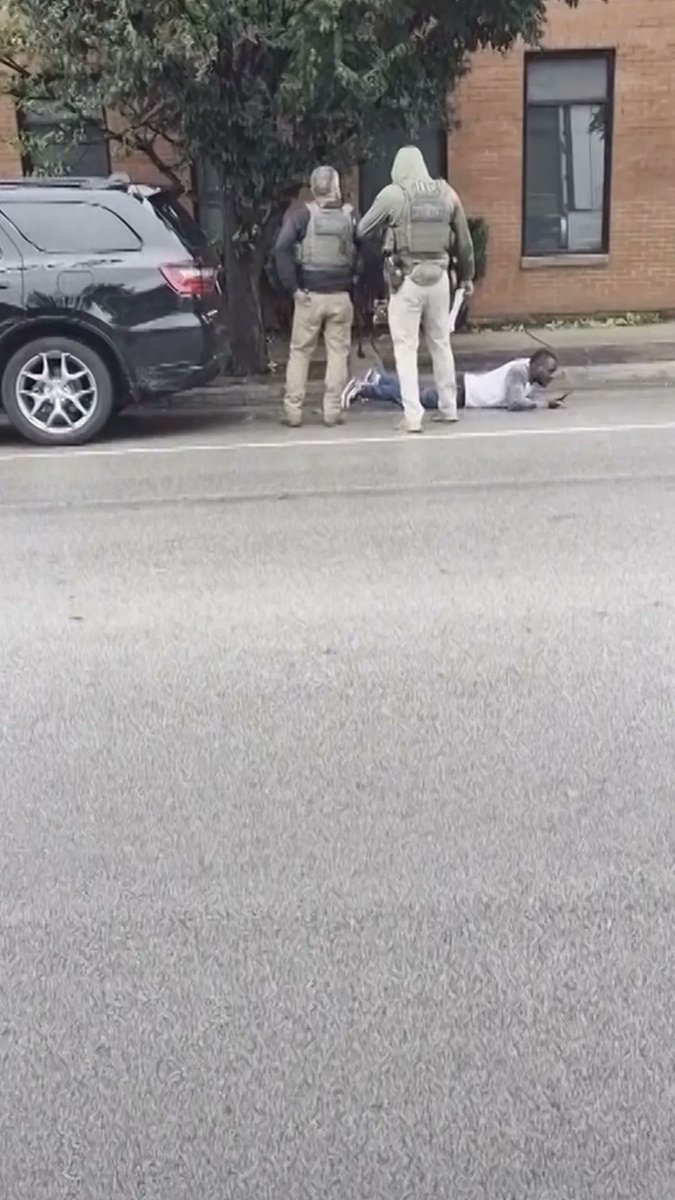 Two armed law enforcement officers in tactical gear stand over a person lying face-down on a street next to a black SUV.