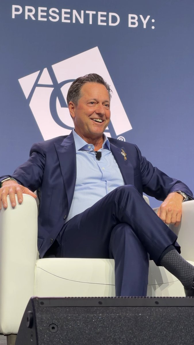 A man in a blue suit and light blue shirt sits smiling on a white chair on stage, with a backdrop reading 'PRESENTED BY:' above a logo featuring the letters 'AC'.